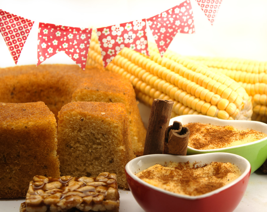 Várias receitas com milho em destaque, como bolo de milho e milho cozido, com bandeirinhas de festa junina ao fundo.